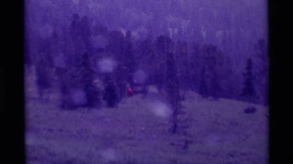 Purple-tinted 8mm frame showing a red raincoat figure far across a Montana meadow with evergreens in the foreground.
