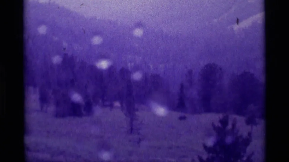 Wide 1975 home-movie view of a Montana meadow with scattered evergreens and distant hikers barely visible.