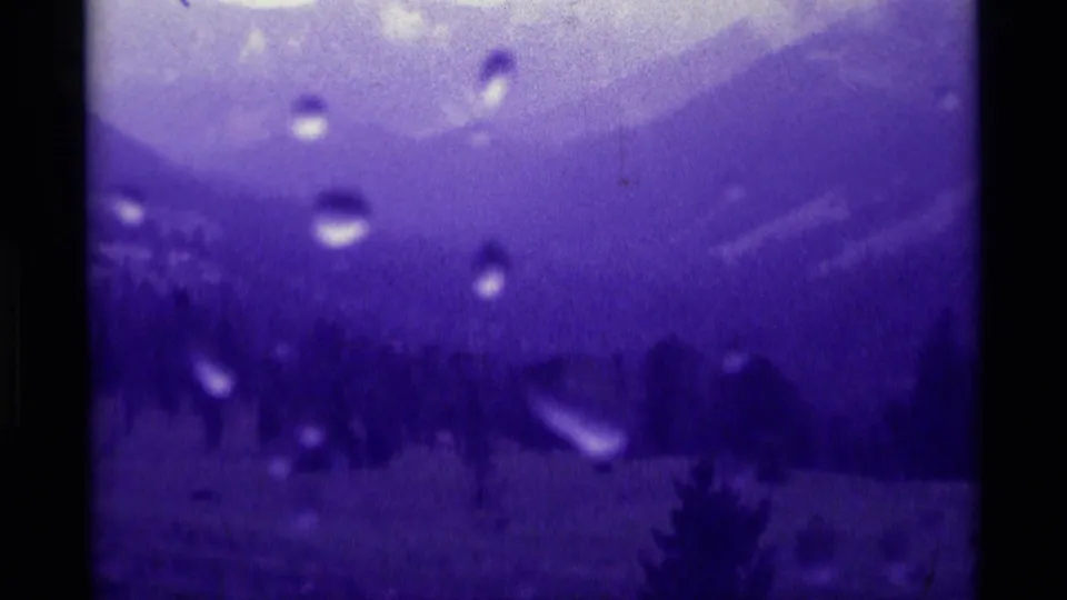 Rain-streaked 1975 8mm view of Montana mountain slopes, dark forest shapes, and lens droplets under a strong purple color shift.