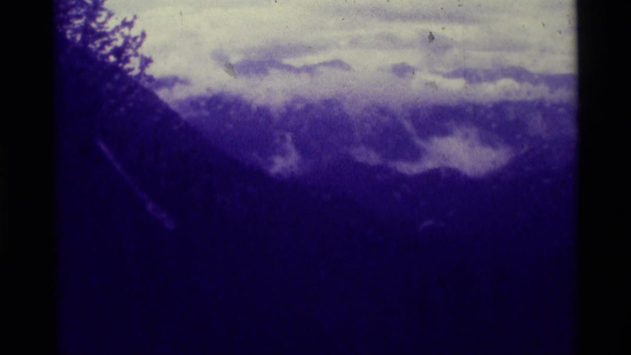 1975 8mm view of a dark Montana mountain landscape with low clouds, pale sky, and heavy purple-blue film tint.