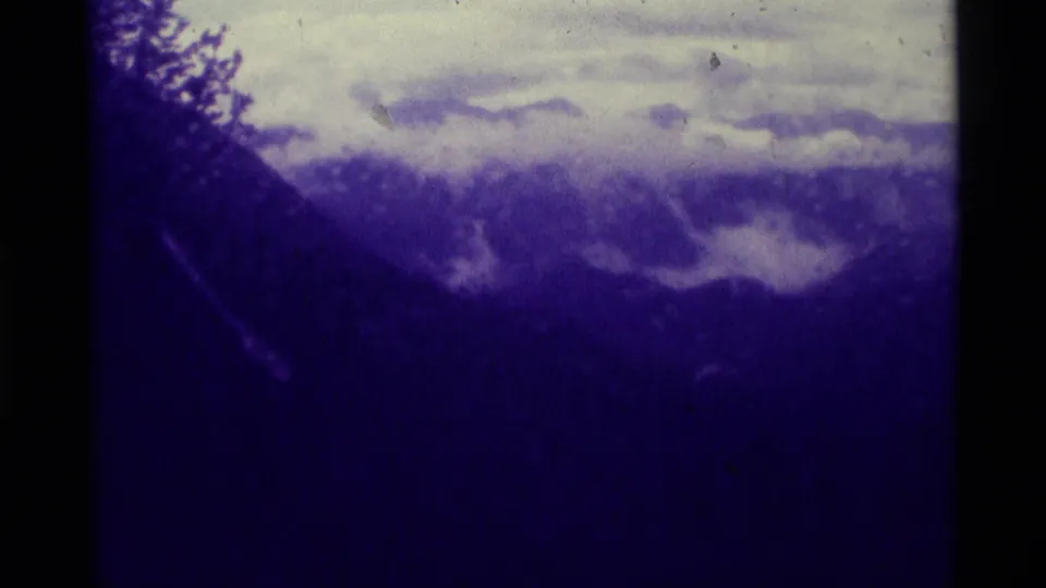 1975 8mm view of a dark Montana mountain landscape with low clouds, pale sky, and heavy purple-blue film tint.