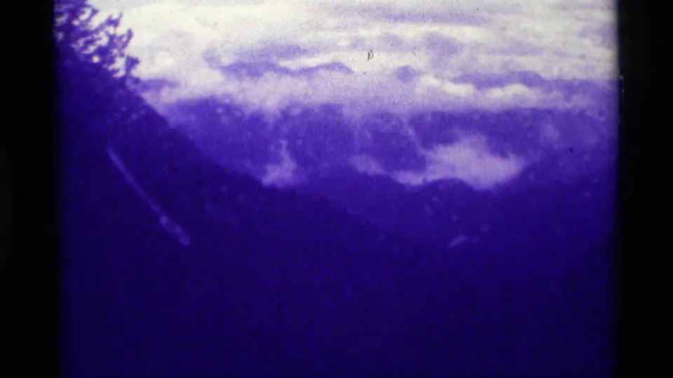 1975 Montana home-movie frame with dark mountain forms, a bright cloud bank, tree silhouette, and visible 8mm grain.