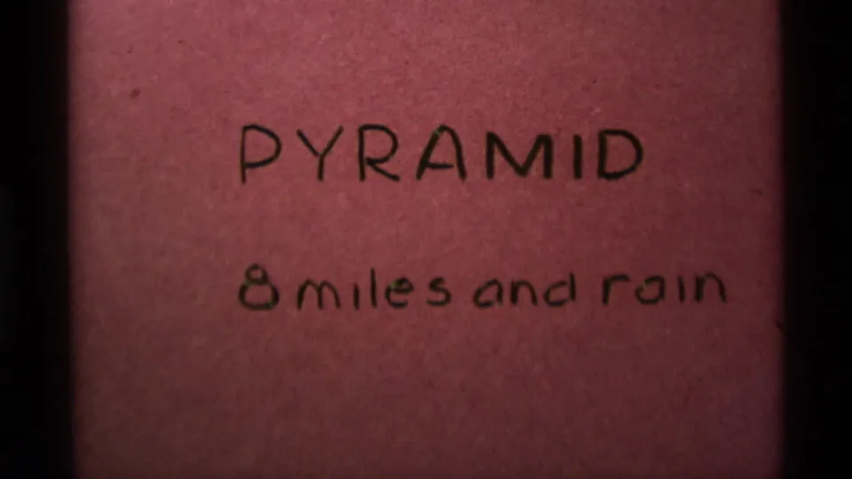 1975 8mm home-movie title card for a Montana hiking segment, handwritten on pink paper with the words Pyramid and 8 miles and rain.
