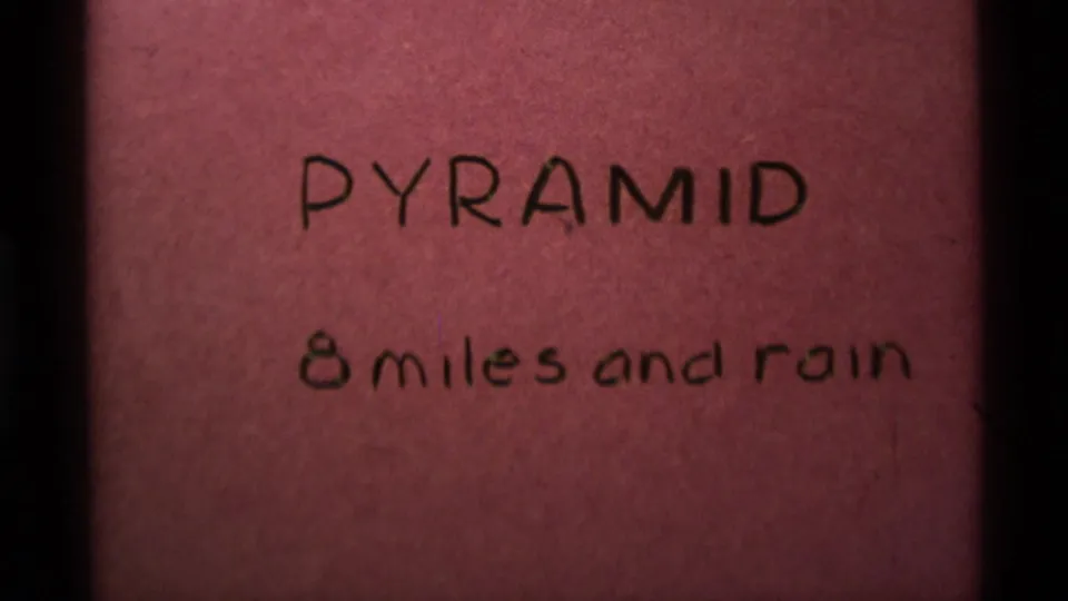 Repeated 1975 8mm title-card frame with Pyramid and 8 miles and rain written on pink paper, framed by dark film edges.