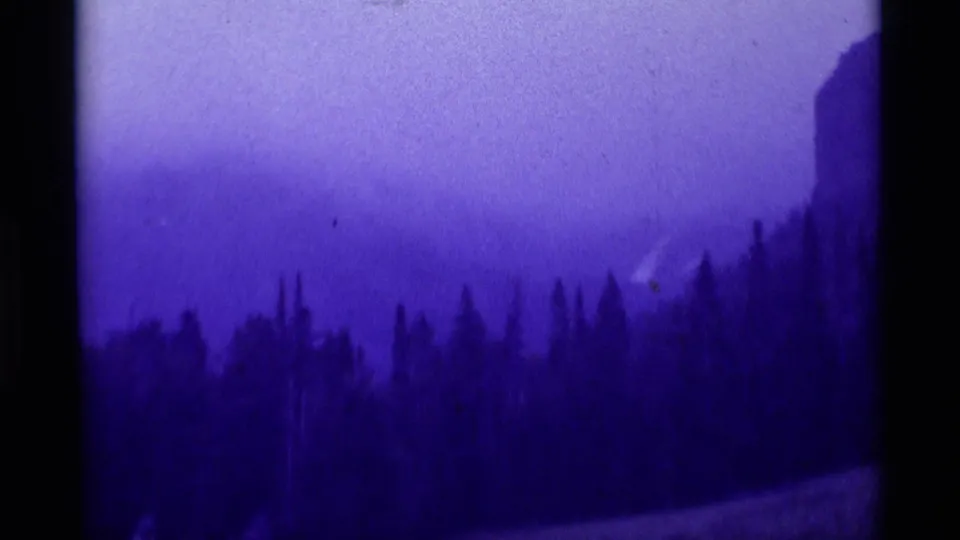1975 8mm frame from Agate Springs area showing a dark evergreen slope, pale sky, and faint distant hikers near the bottom edge.