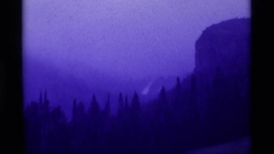 1975 8mm Agate Springs, Montana view of evergreens below a misty mountainside with a cliff-like rise at right.