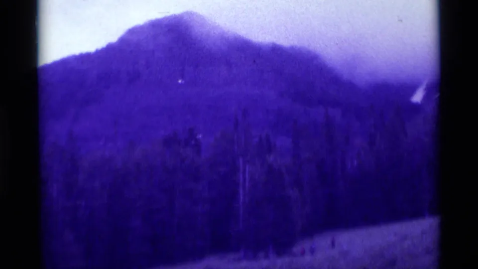 1975 8mm Agate Springs, Montana frame showing a misty mountain ridge, evergreen band, and pale ground with tiny distant figures.