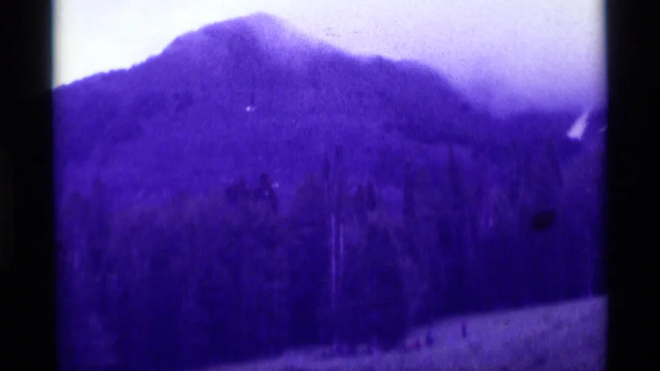 1975 8mm Agate Springs, Montana frame of a forested mountain under haze, with small distant figures on the lower slope.