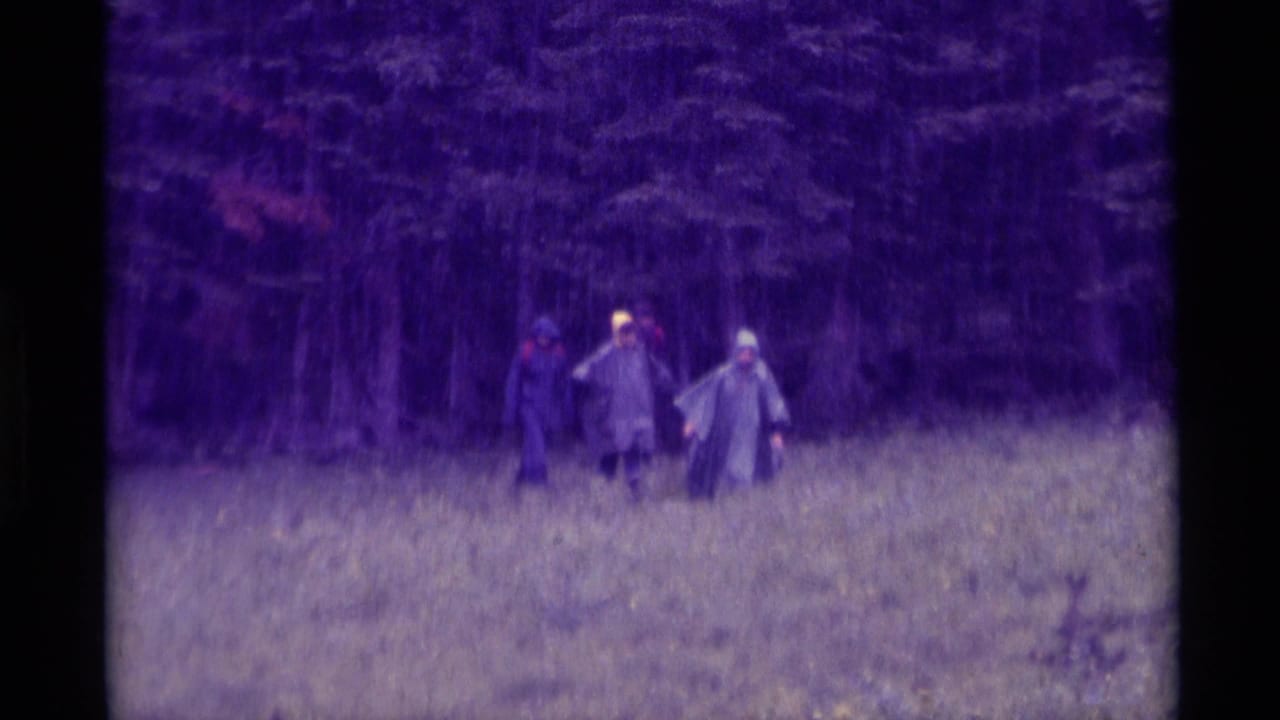 1975 8mm home-movie frame from Agate Springs, Montana, showing distant people in rain gear crossing wet grass before trees.