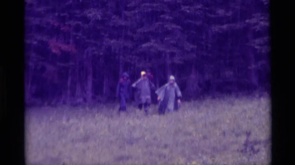 1975 8mm home-movie frame from Agate Springs, Montana, showing distant people in rain gear crossing wet grass before trees.