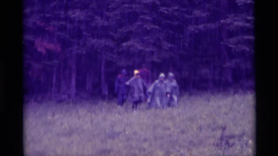 Distant figures in hooded ponchos move through a rainy field in supported 1975 Agate Springs, Montana home-movie footage.