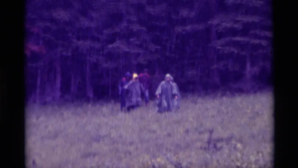 A soft 8mm frame shows rain-coated walkers in a Montana field, with a dark wooded background and visible color fading.
