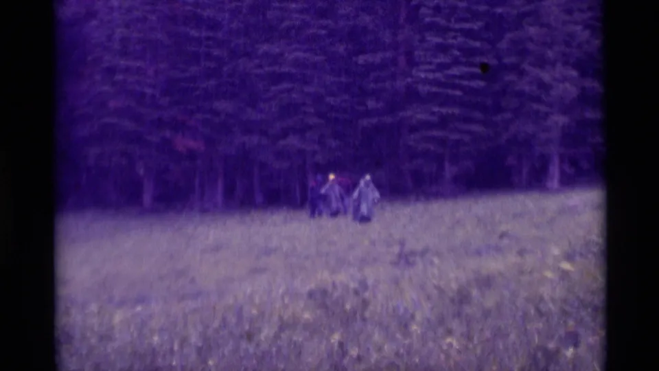 Wide 1975 8mm view of distant poncho-clad figures crossing a wet field near forest in Agate Springs, Montana.