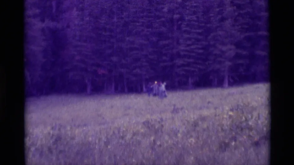 1975 Agate Springs, Montana home-movie frame with small distant walkers crossing a meadow beneath dense evergreens.