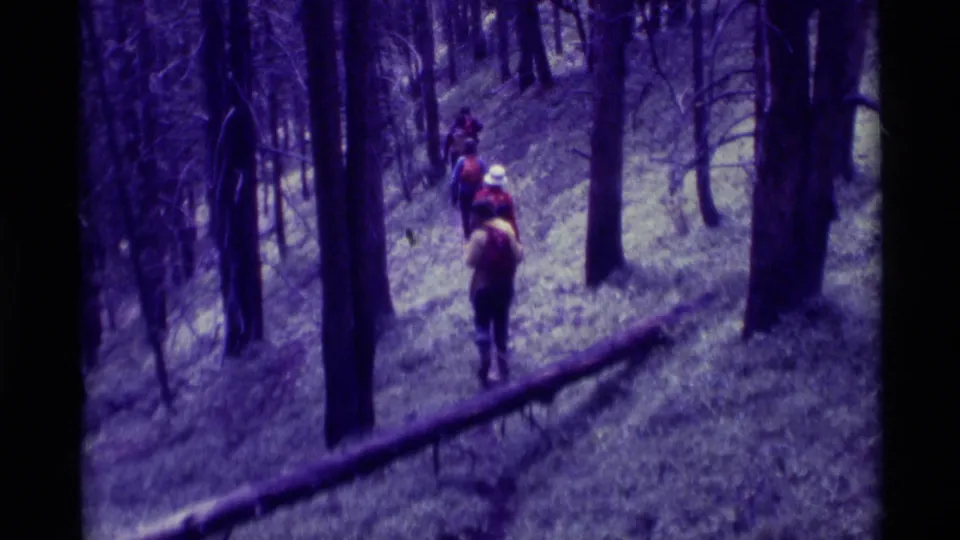 Distant hikers, including one with a white hat, climb a forested hillside in 1975 Agate Springs, Montana home-movie footage.