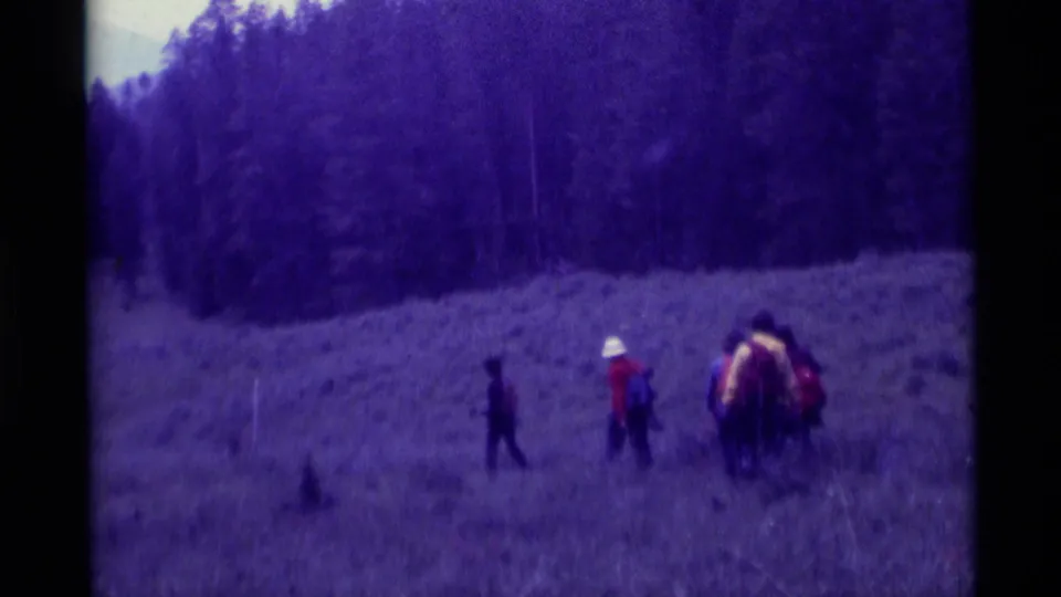 Several hikers cross a grassy meadow in Agate Springs, Montana, with a forested slope behind them.