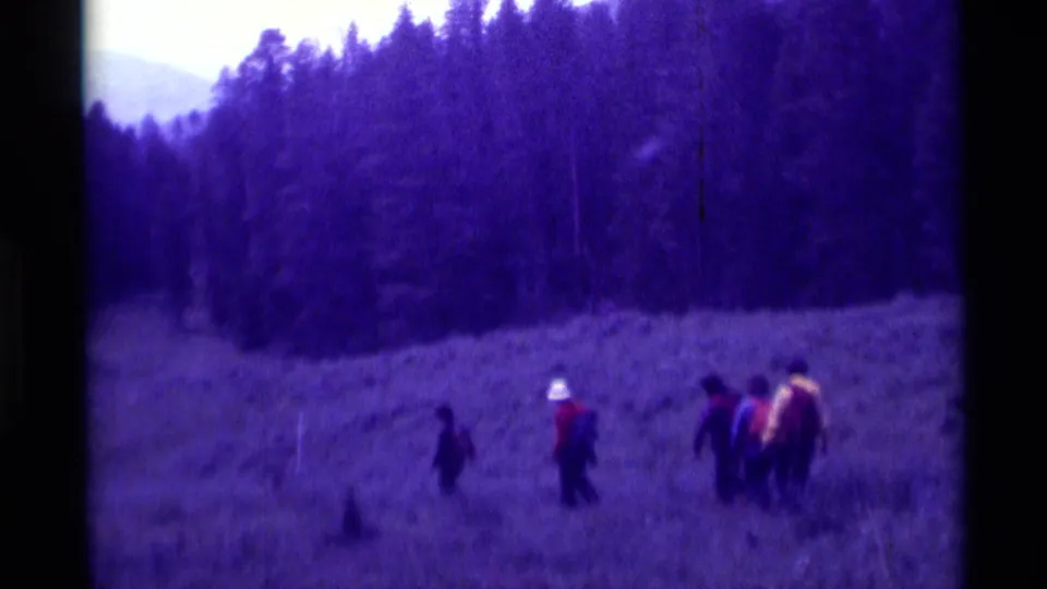 The hiking group continues across the meadow in 1975 Montana, with soft-focus trees and distant slope in the background.