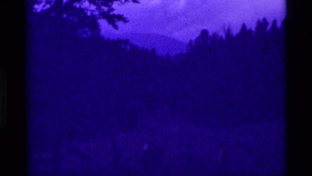 1975 8mm frame from Agate Springs, Montana, showing distant hikers in a forested mountain clearing under a strong purple color cast.