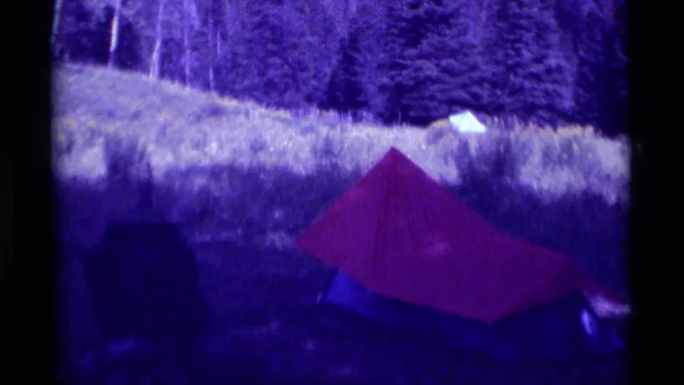 Closer 8mm view of a red tent with blue lower fabric in a grassy Agate Springs field, with trees behind it.