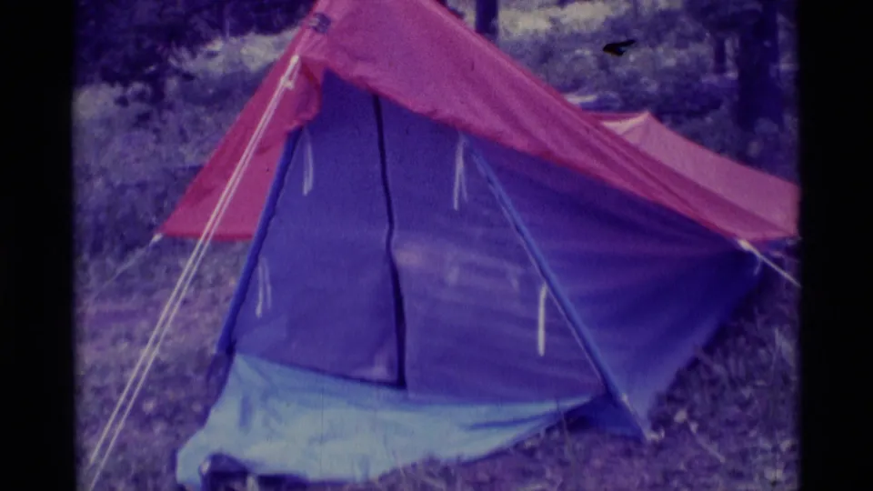 1975 8mm camping frame showing the blue tent entrance beginning to open beneath the red fly in the woods.