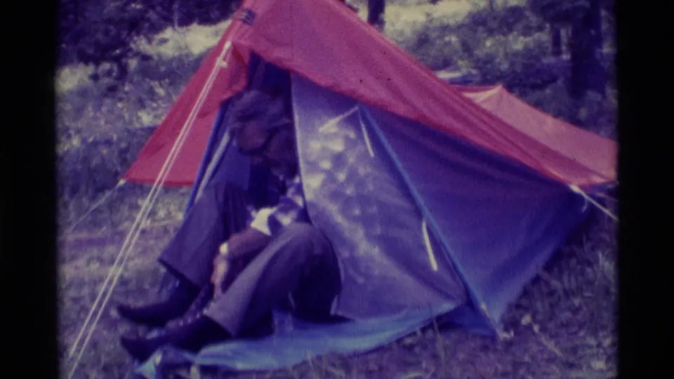 The 1975 8mm frame shows the adult camper seated at the tent entrance in a wooded Montana campsite.
