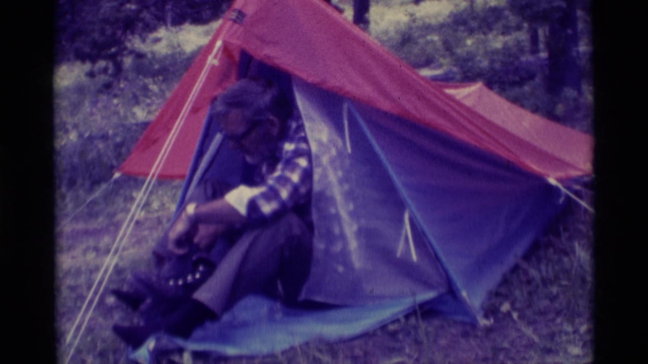 In 1975 Montana home-movie footage, an older man in glasses and a plaid shirt sits at a red and blue tent entrance adjusting a boot.
