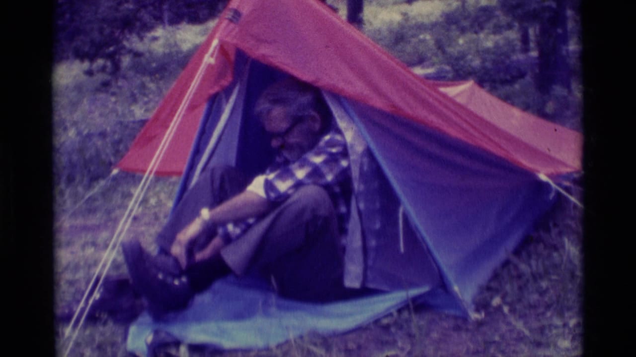 Older man seated inside a red and blue tent, pulling at a boot near the entrance.