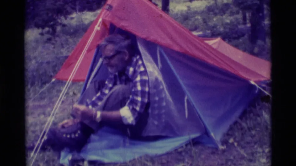 At the wooded Montana campsite, the older man bends over his boot beside the red and blue tent in soft 8mm color.