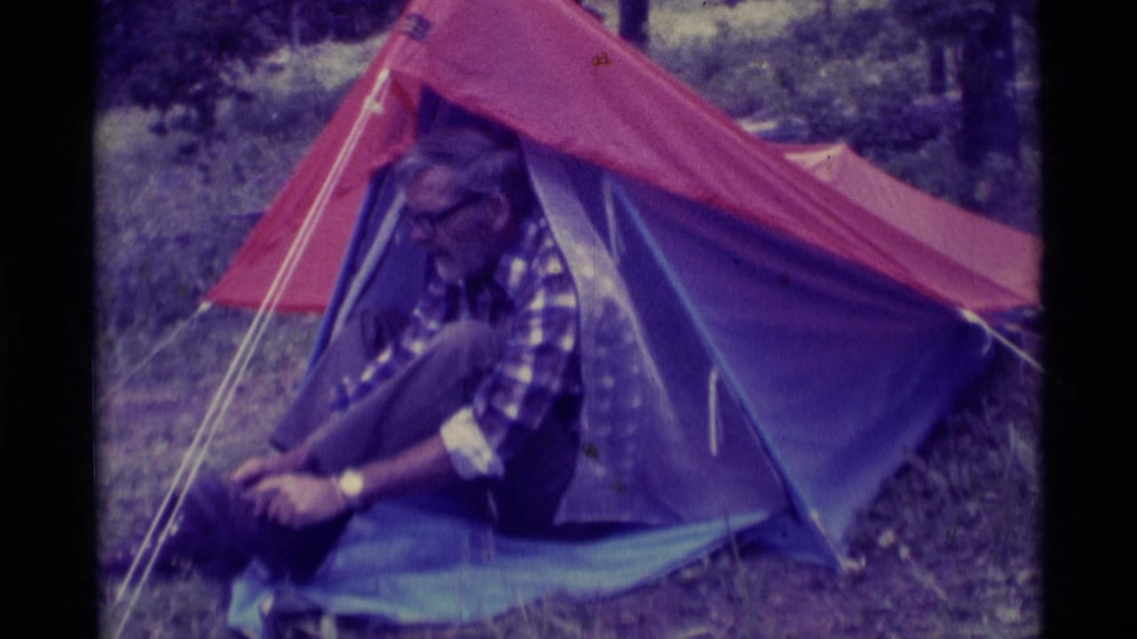 In 1975 Montana home-movie footage, an older man in a plaid shirt sits at a tent entrance and adjusts a hiking boot.