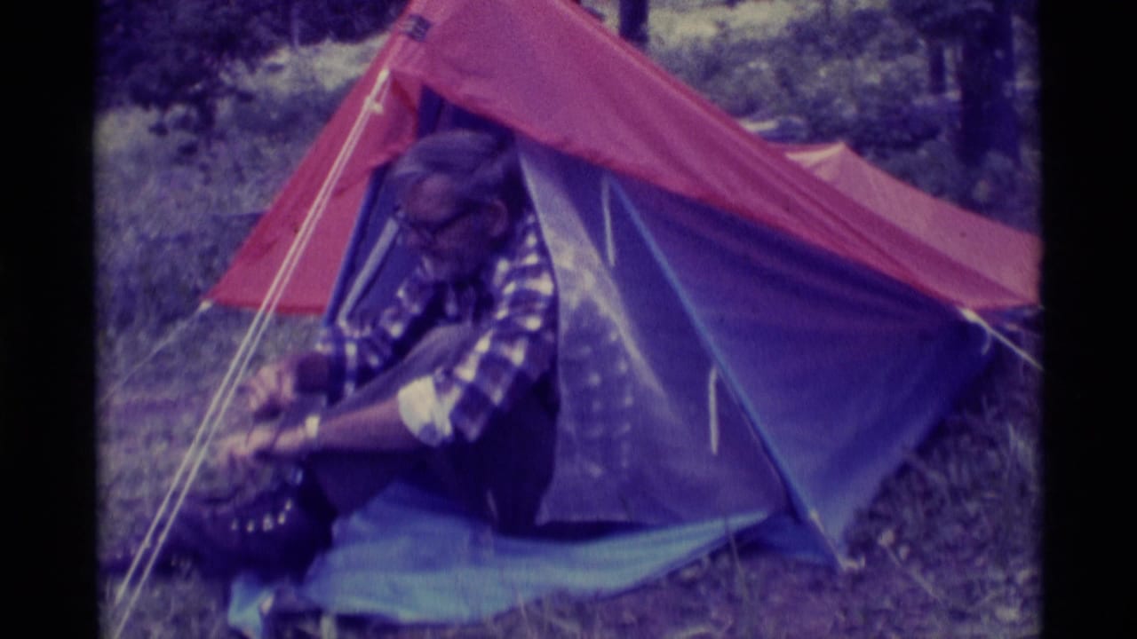 Older man in a plaid shirt seated in front of a small tent, working on his boot.