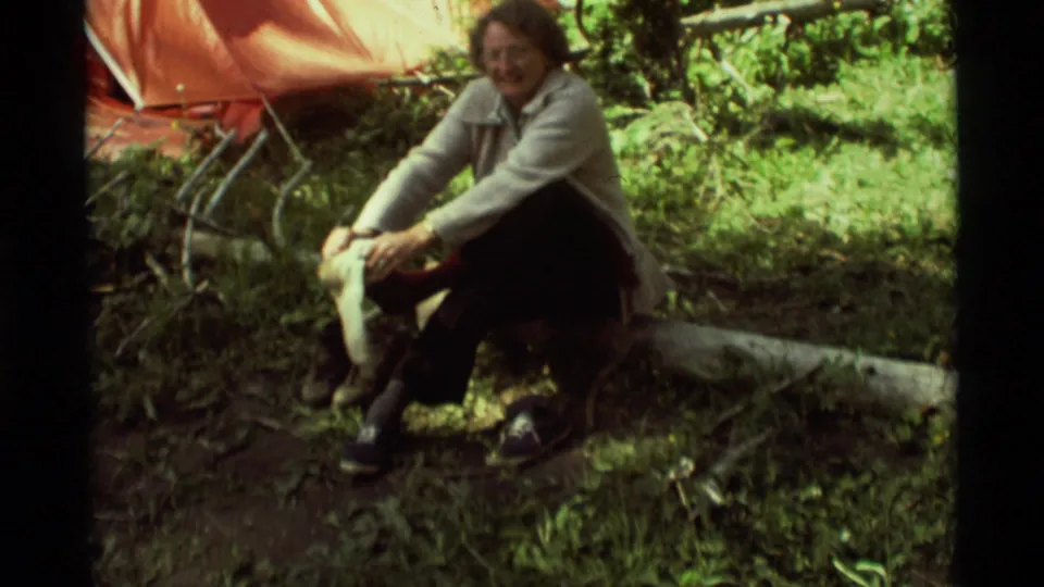 An older woman in a light jacket sits on a log near the tent and adjusts footwear in the forest campsite.