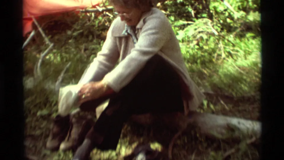 The older woman leans forward over her footwear on a log at the 1975 Agate Springs campsite, with green forest behind her.