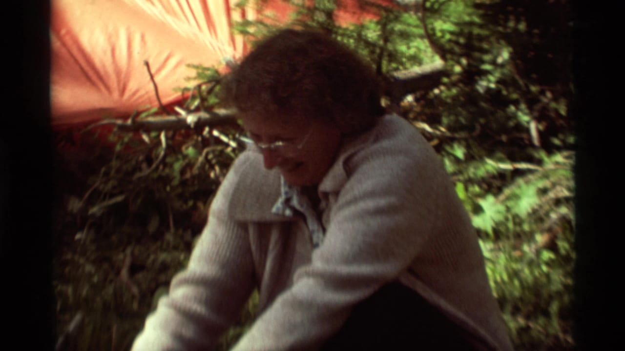 In 1975 Montana home-movie footage, a woman with glasses crouches in a wooded campsite near orange tent fabric.
