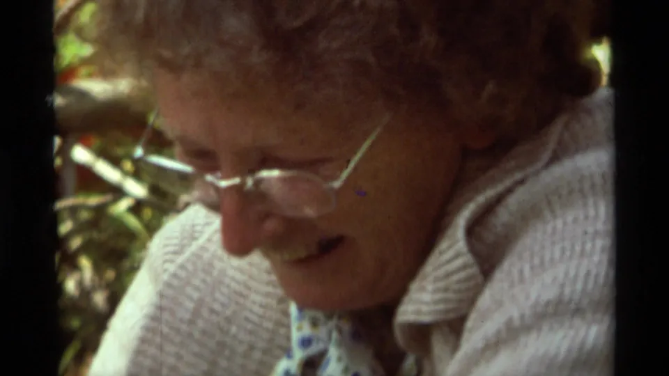 The 1975 Montana home-movie frame shows the woman leaning over the campsite task with a slight smile visible.