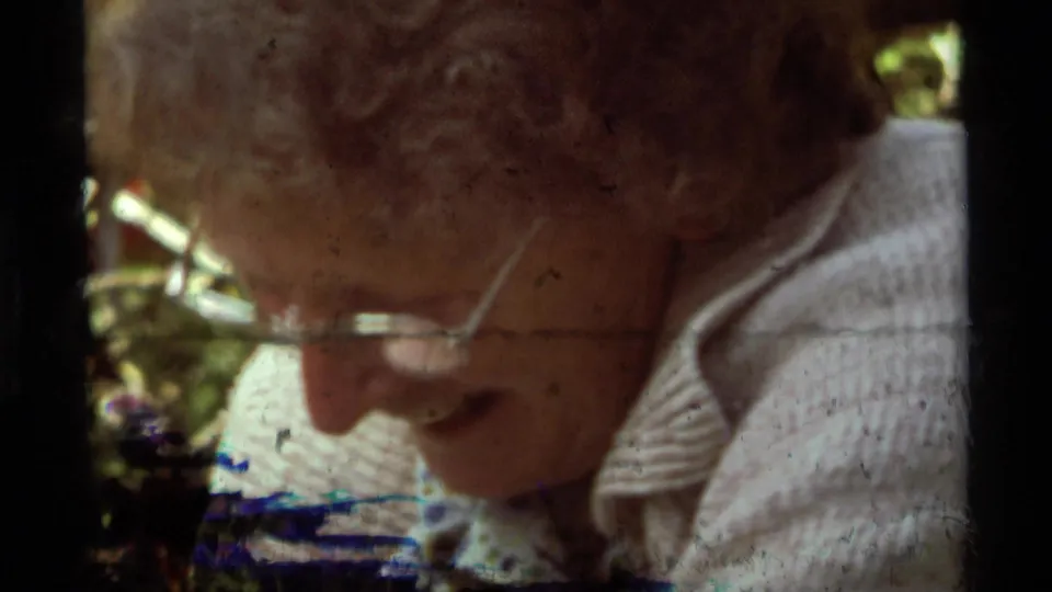 Close 1975 8mm frame from Agate Springs, Montana, showing an older person in glasses near the camera with campsite color behind them.
