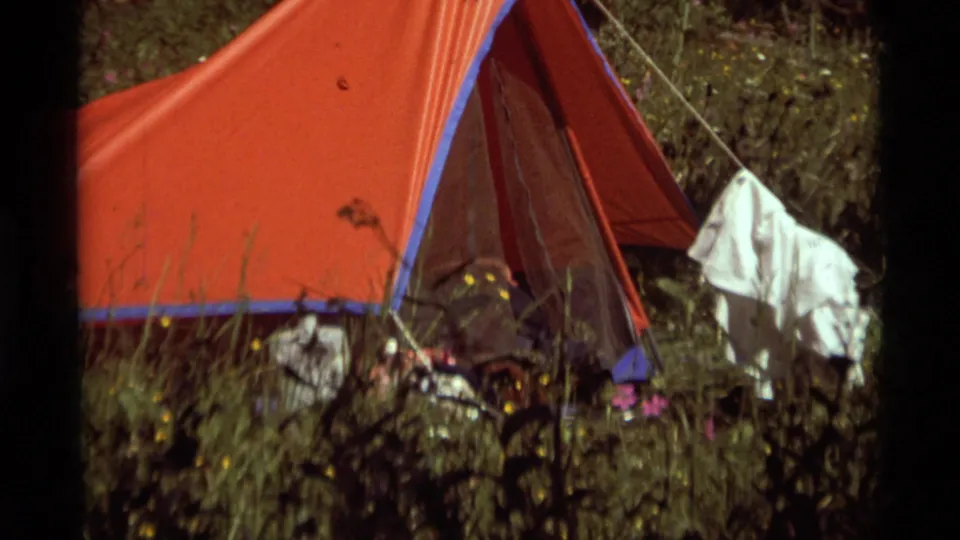 Agate Springs, Montana, 1975 campsite frame with a red tent, white clothing on a line, grass, flowers, and camping gear in view.
