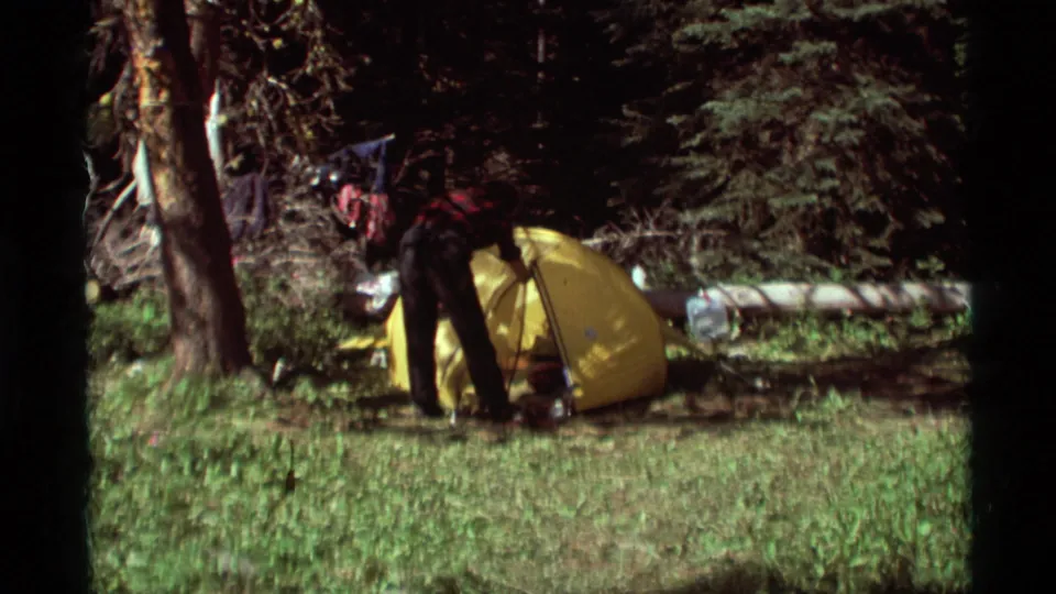 Person in a red plaid shirt and dark pants working beside a yellow tent in a wooded campsite clearing in 1975 Montana footage.