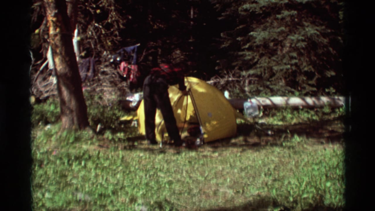 Camper stands beside a yellow tent in a wooded clearing.