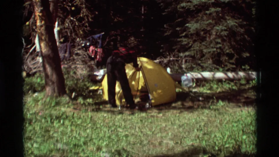 In 1975 Agate Springs, Montana footage, a camper in plaid stands by a yellow tent among trees and grass.