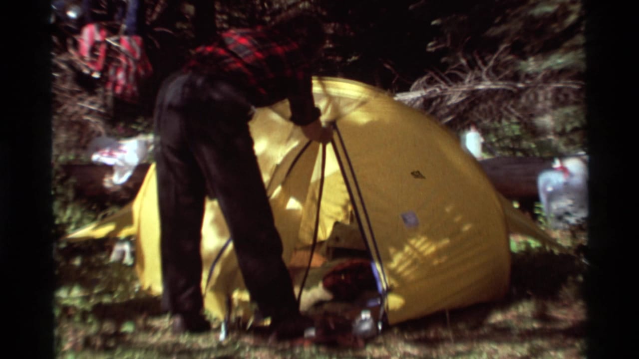 The camper leans over a yellow dome tent, with metal poles and camping containers visible in the wooded Montana setting.