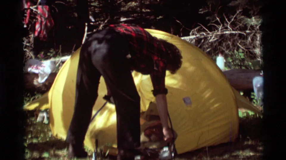 A plaid-shirted camper reaches toward gear or a fastening point near the yellow tent entrance in 1975 home-movie footage.