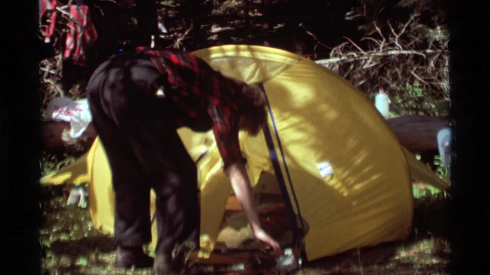 The camper bends beside the yellow tent under dappled forest light, with poles and containers around the campsite.