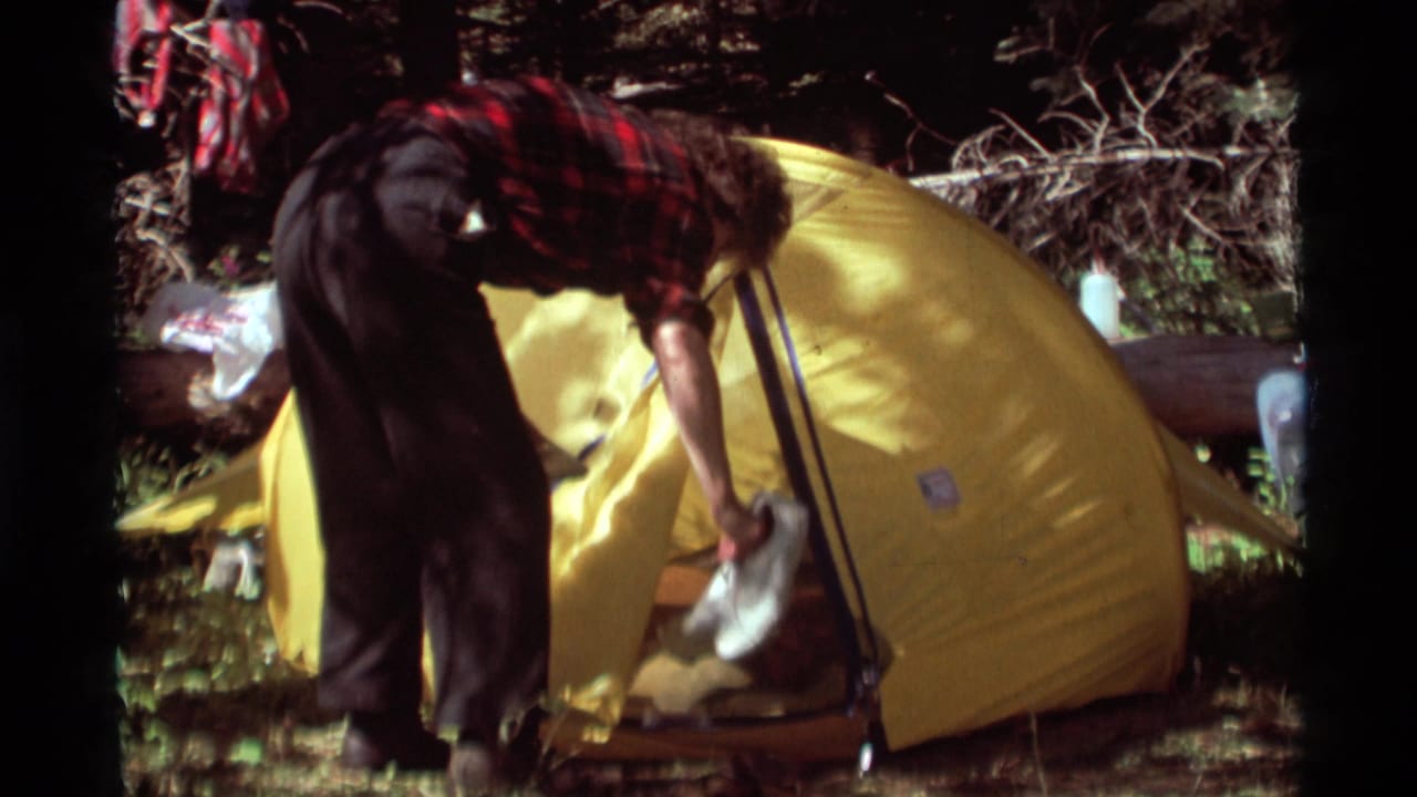 In 1975 Agate Springs, Montana footage, a camper bends near the entrance of a yellow tent among trees and campsite gear.
