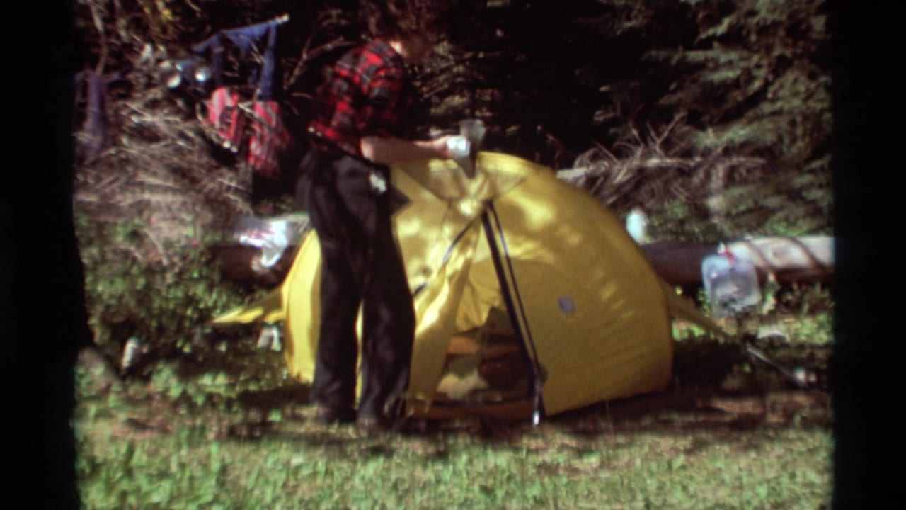 Camper stands over a yellow tent with poles and gear in a shaded forest clearing.