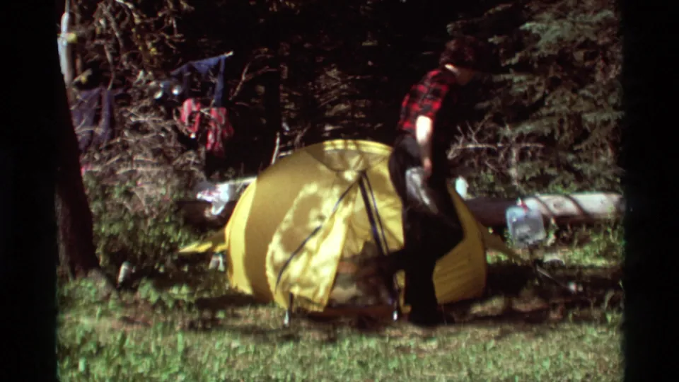 A camper stands next to the yellow tent in the Montana campsite, with trees, grass, and hanging gear visible around him.