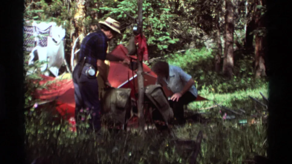 Two adult campers, one wearing a straw hat, assemble a red tent in a grassy Agate Springs forest campsite.