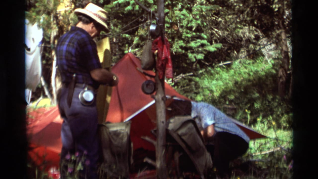 Standing camper faces a red tent while a second adult bends near gear in the grass.