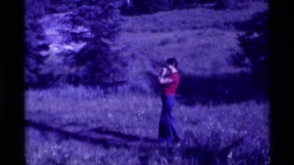 Purple-shifted 8mm frame showing a person in a red shirt standing alone in a grassy field, likely from the same reel.