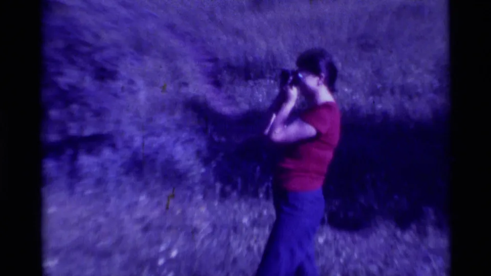 In 1975 Agate Springs, Montana home-movie footage, a woman in a red shirt and blue pants looks through a scope outdoors.