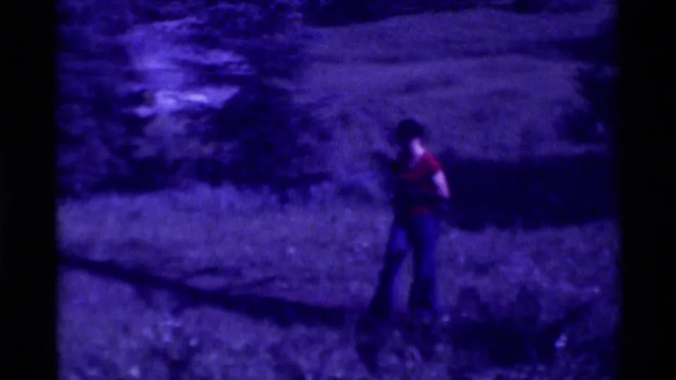 A wider 1975 home-movie view shows the woman standing in grass, with trees and shadowed ground around her.